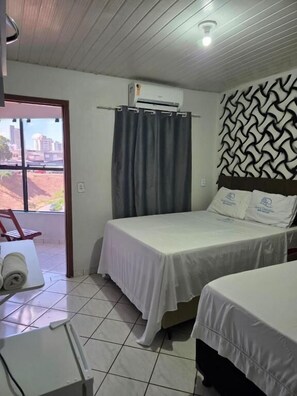 Quarto de hotel com duas camas, janela com cortinas, ar condicionado e vista para edifícios.