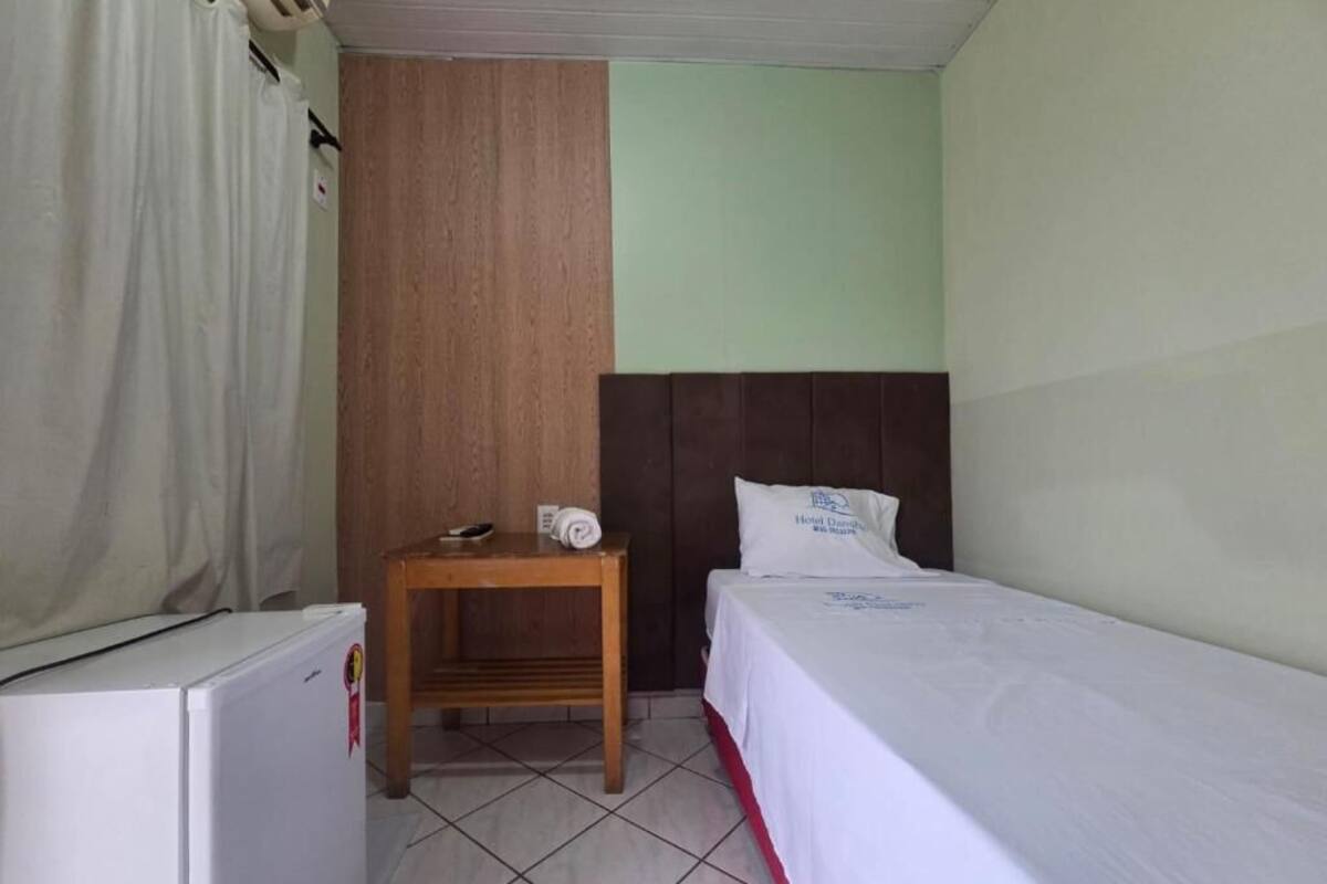 Una habitación de hotel pequeña con una cama individual, una mesita de noche de madera, un refrigerador blanco y una cortina.