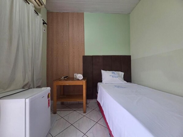 Una habitación de hotel pequeña con una cama individual, una mesita de noche de madera, un refrigerador blanco y una cortina.