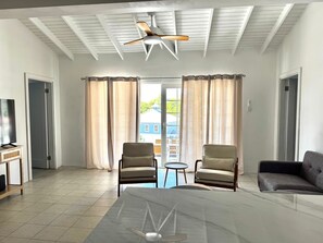 Una habitación amplia con ventilador de techo, dos sillas, un sofá, un televisor y vistas a una casa azul a través de las cortinas.