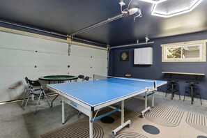 Ein Tischtennistisch, Stühle, ein Dartspiel und eine Dartscheibe in einem Raum mit einer Deckenleuchte.