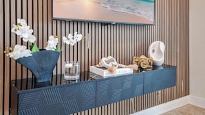 Una sala de estar moderna con un televisor grande montado en una pared de madera, una mesita consola azul con objetos decorativos y orquídeas blancas en un jarrón.