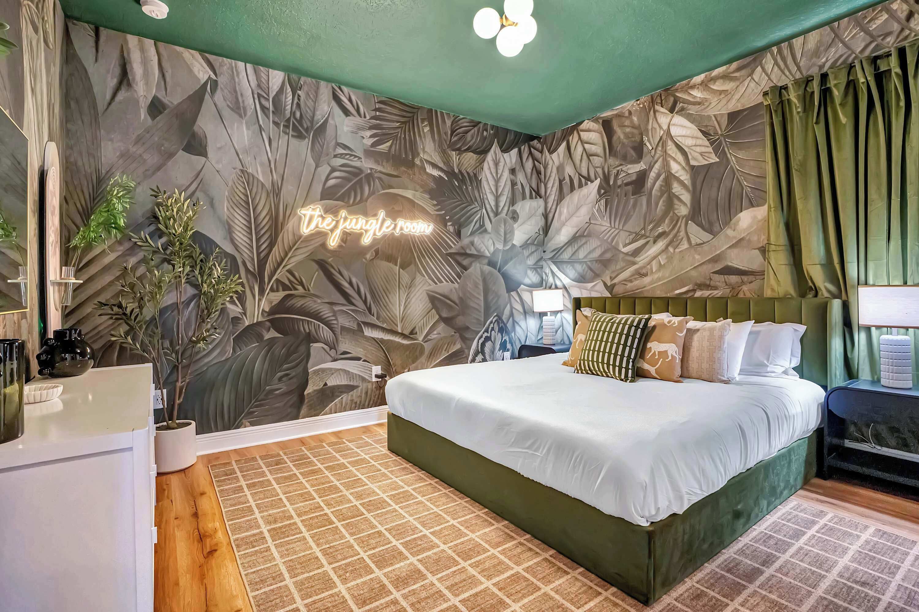 A bedroom with a bed, a nightstand, a lamp, a potted plant, and a neon sign.
