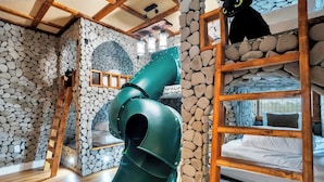 Una habitación con pared de piedra, una cama litera, un tobogán verde y una escalera de madera.