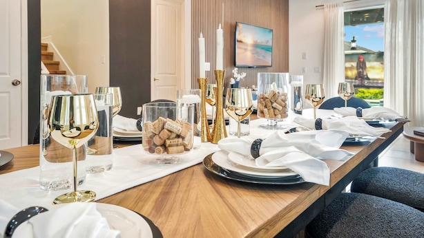 Una mesa de comedor preparada con copas de vino, tapones de corcho y elementos decorativos, con un televisor montado en la pared y una ventana con cortinas.