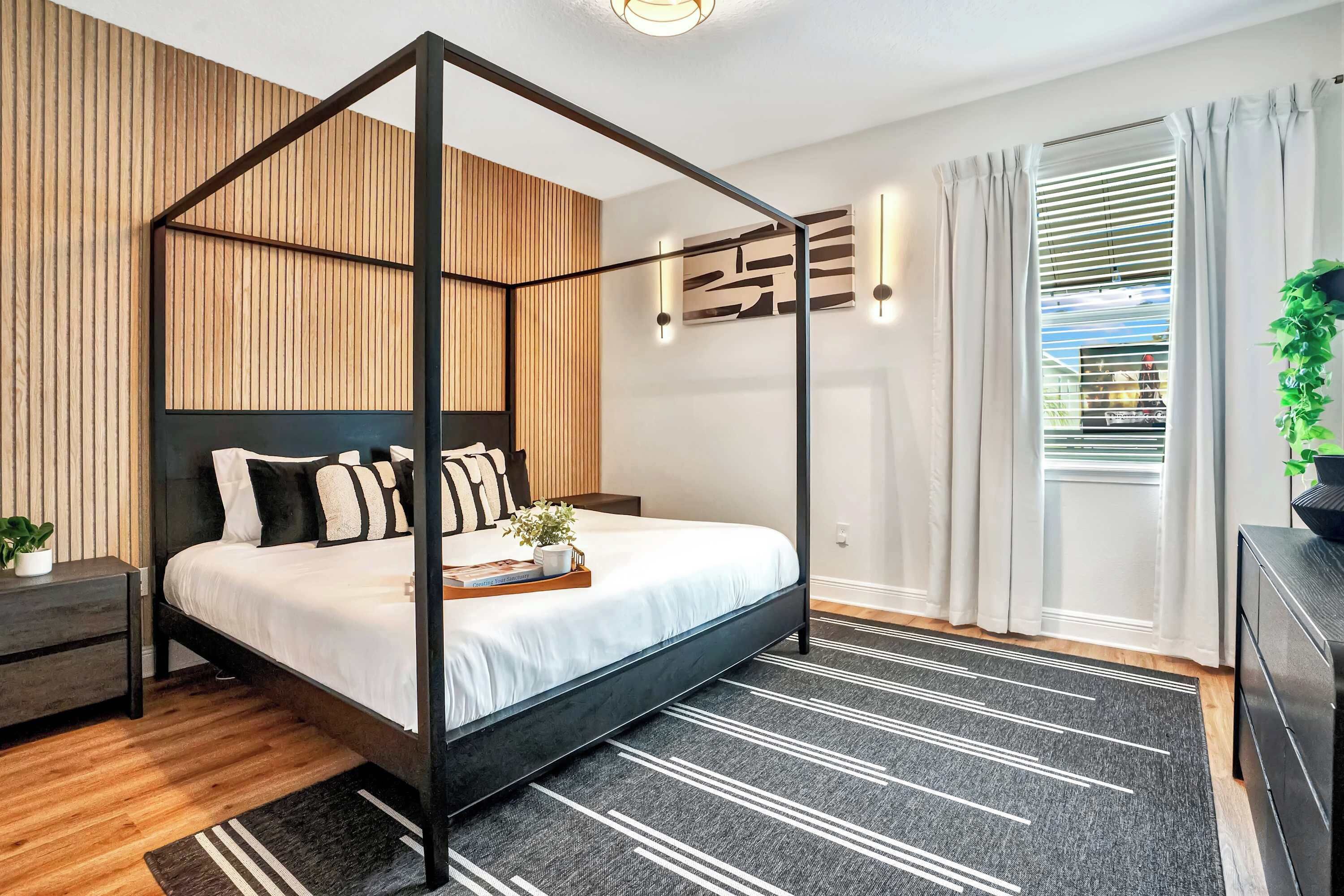 Un dormitorio moderno con una cama con dosel, un panel de pared de listones de madera, estructura metálica negra, alfombra oscura y una mesita de noche con una planta.