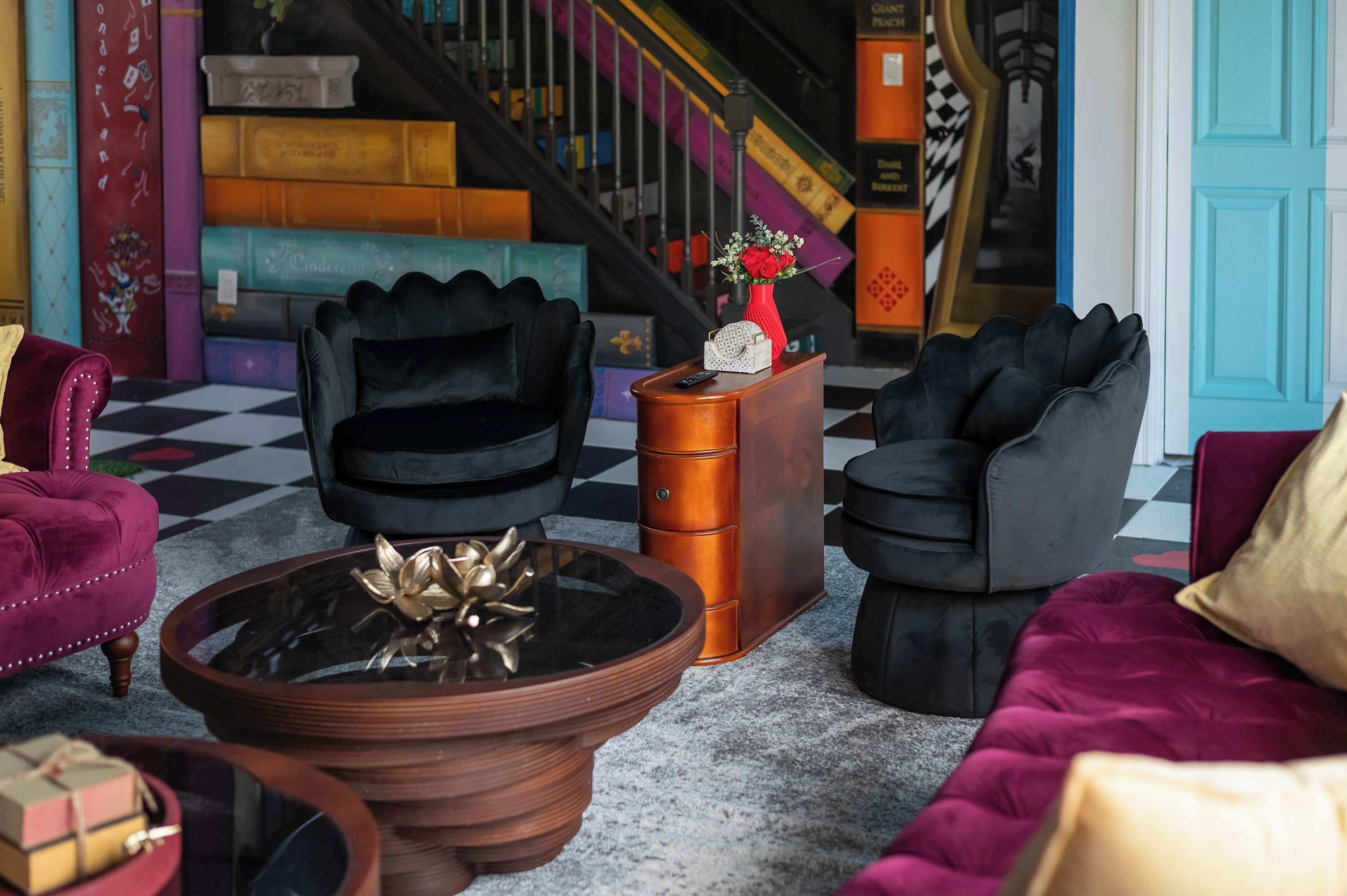Een geruite vloer, een zwarte fauteuil, een rode fauteuil, een houten bijzettafeltje met een vaas, en een trap met kleurrijke treden.