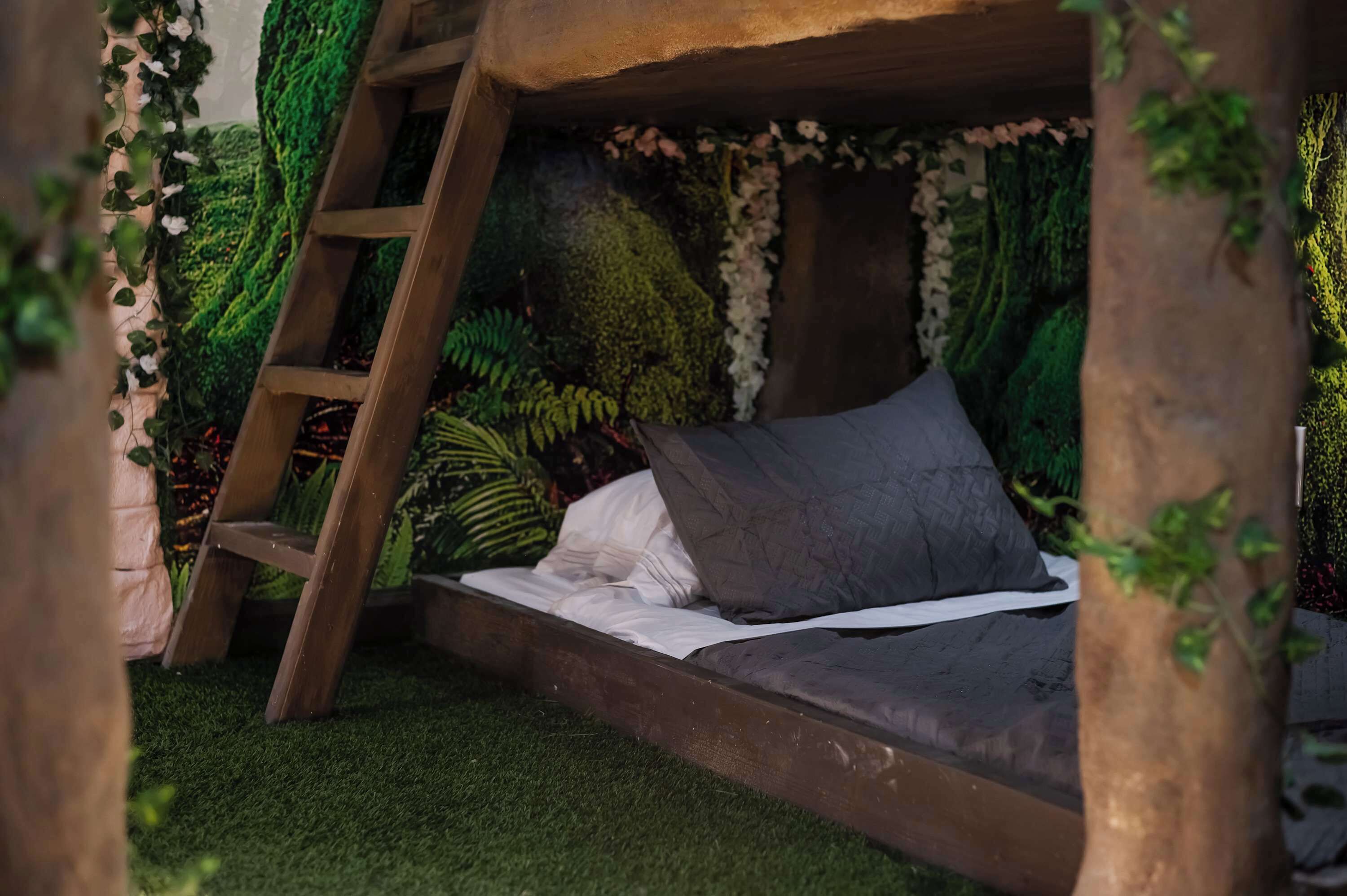 Een rustiek houten bed met een ladder, omgeven door weelderige begroeiing en kunstmos.