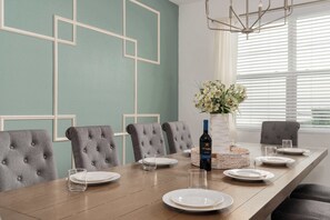 A dining table set with plates, glasses, and a bottle of wine, surrounded by tufted chairs in a room with geometric wall designs and a window with blinds. - Family retreat w/ theater, pool & hot tub (Kissimmee)