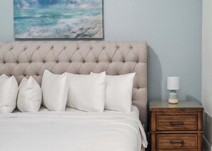 Un lit doté d’une tête de lit capitonnée, de draps blancs et d’une lampe de chevet posée sur une table de nuit en bois.