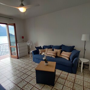 Ein Wohnzimmer mit einem blauen Sofa, einem Holztisch, einer weißen Lampe, einem Deckenventilator und Meerblick durch eine Schiebetür.