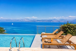 Ein Poolbereich mit Liegestühlen, Blick auf das Meer und ein Segelboot in der Ferne.