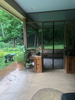 Ein überdachter Patio-Bereich mit einer Holzbank, Kübelpflanzen und einem Gewächshaus.