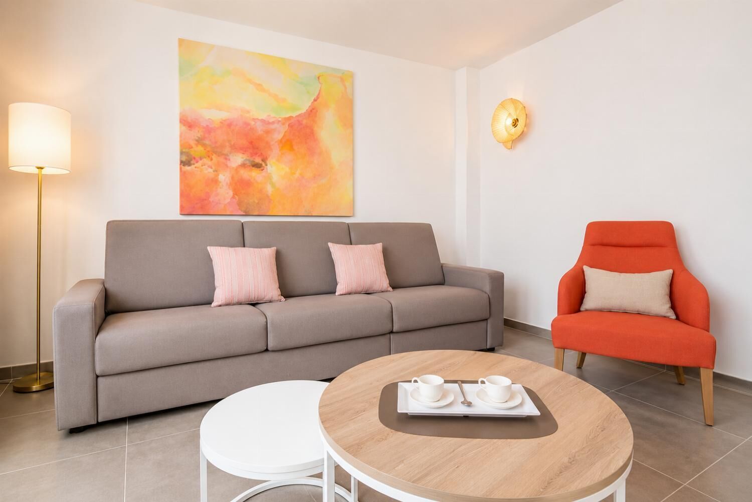 Ein modernes Wohnzimmer mit grauem Sofa, einem runden Couchtisch aus Holz, einem weißen Beistelltisch, einem roten Sessel, einer Stehlampe und einer Wandleuchte.