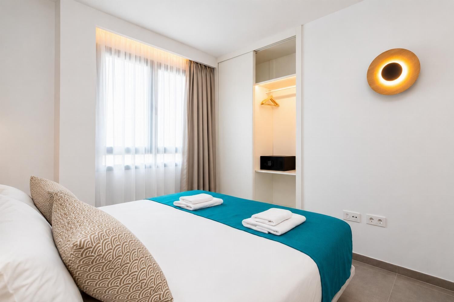 A hotel room with a bed, a bedside table, a wardrobe, and a wall-mounted light fixture.