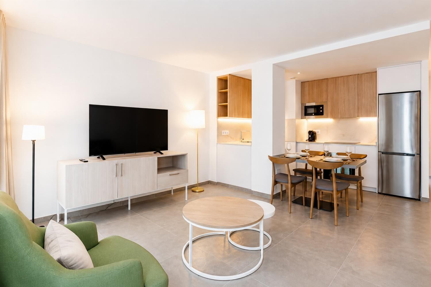 A modern living room with a flat-screen TV, a green sofa, a round coffee table, and a dining area with wooden chairs and a small table.