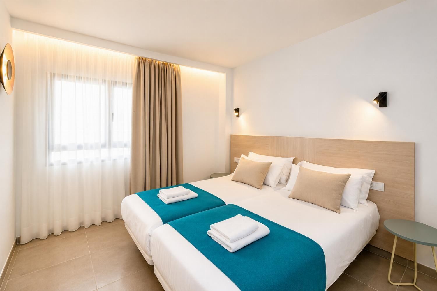 Een hotelkamer met twee bedden, een groot raam met gordijnen, een wandlamp en een klein tafeltje.