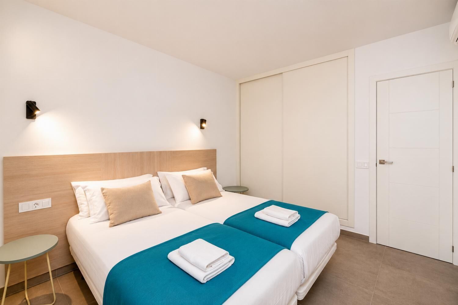 A hotel room with a double bed, a small side table, a wall-mounted light, and a door.