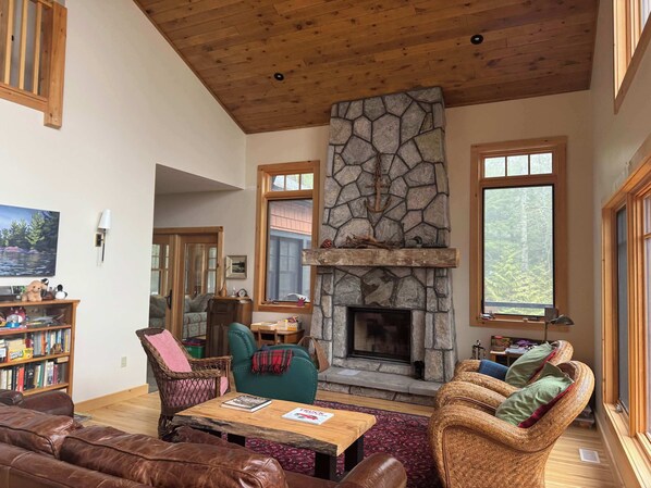 A cozy living room with a stone fireplace, wooden ceiling, and large windows.