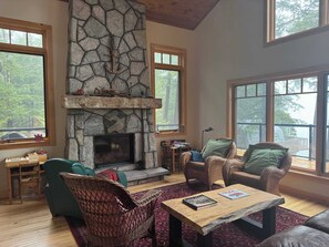 A spacious living room with a stone fireplace, large windows, wicker furniture, and a wooden coffee table.