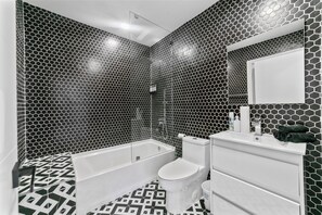 Ein Badezimmer mit sechseckigem Fliesenmuster, einer weißen Badewanne, einer Toilette und einem weißen Waschtisch mit Spiegel.