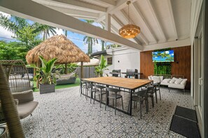 A patio with a wooden table and chairs, a thatched-roof structure, and a TV screen displaying a tropical scene.