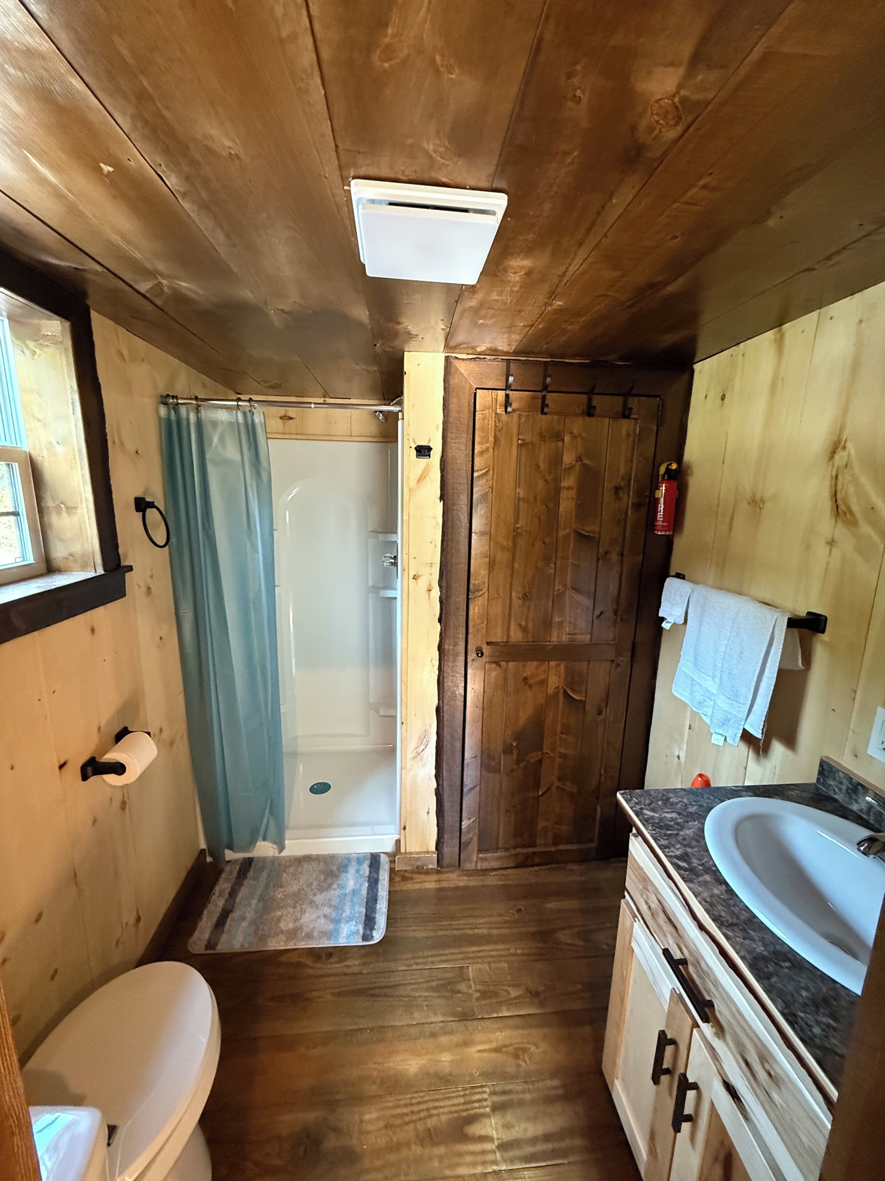 Une salle de bain rustique, avec des murs en bois, comprenant une douche, des toilettes, un lavabo et une porte donnant sur une autre pièce.