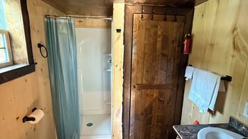 Une salle de bain rustique, avec des murs en bois, comprenant une douche, des toilettes, un lavabo et une porte donnant sur une autre pièce.