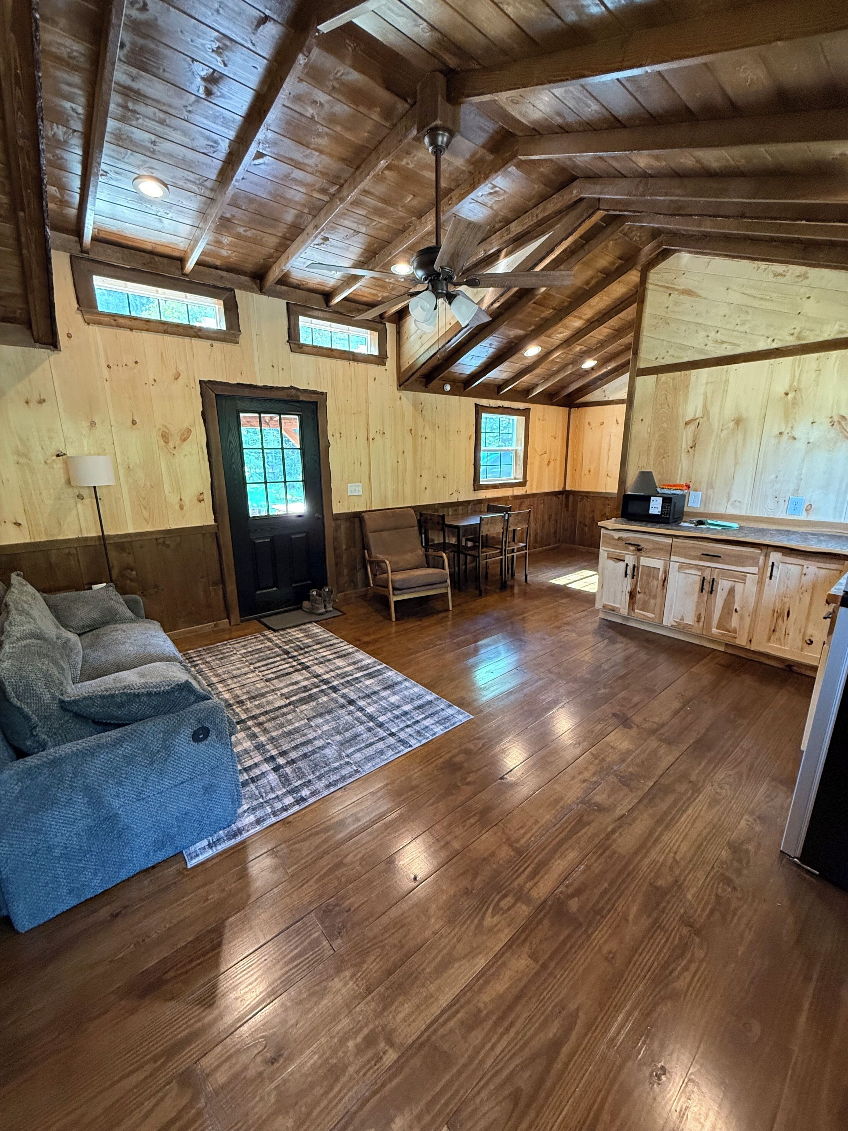 Une pièce spacieuse dotée de planchers en bois, d’un ventilateur de plafond, d’un coin repas avec des chaises, d’un canapé et d’un coin cuisine avec des placards.