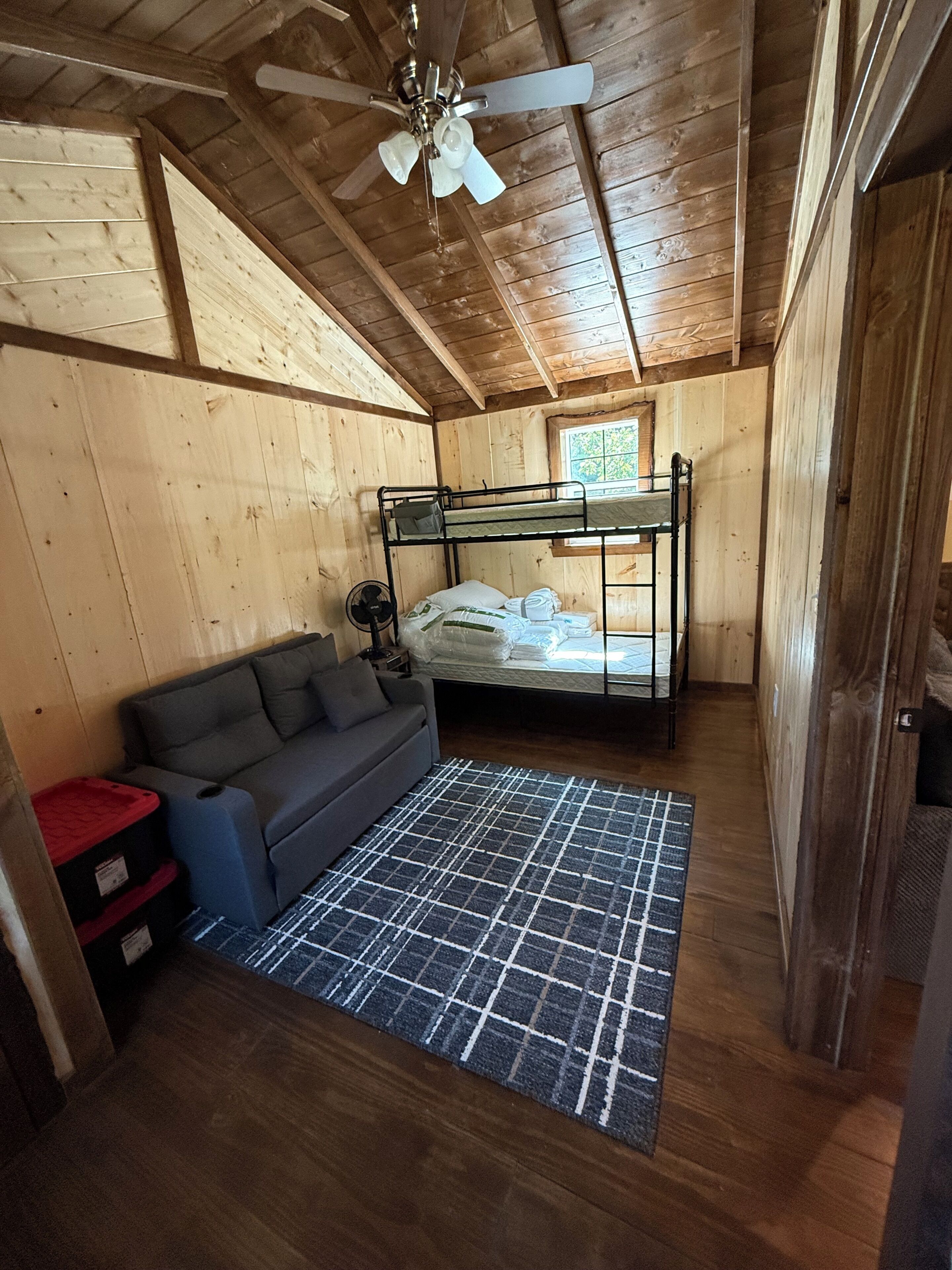 L’intérieur d’une cabane en bois, avec des lits superposés, un canapé, un ventilateur et une fenêtre.
