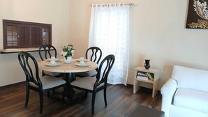 Uma sala de jantar com uma mesa redonda, quatro cadeiras, um vaso com flores, uma mesinha lateral com livros e um sofá branco.
