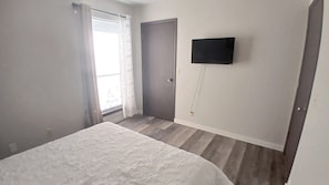 Une chambre à coucher avec un lit, une porte, une fenêtre avec des rideaux et un téléviseur fixé au mur.