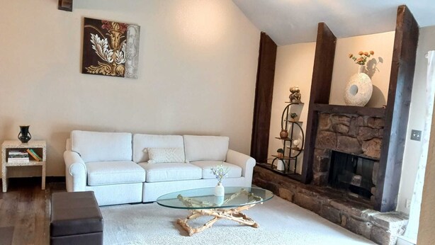 Sala de estar com sofá branco, mesa de centro de vidro, lareira e prateleira decorativa.
