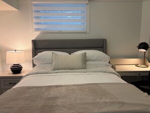 A neatly made bed with a grey headboard, a bedside lamp, and a window with blinds. - New, Cozy 1-Bed, Bsm Suite. with AC in Beautiful Edmonton, Prívate Entry. (Edmonton)