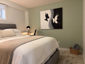 Ein Schlafzimmer mit Bett, Schreibtisch, Lampe, Wandbild, Fenster und Korb.