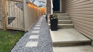 A pathway leading to a house with a wooden fence, a stone pillar, and a flower planter on the steps. - New, Cozy 1-Bed Room, Bsm Suite. with AC in Beautiful Edmonton, Prívate Entry. (Edmonton)
