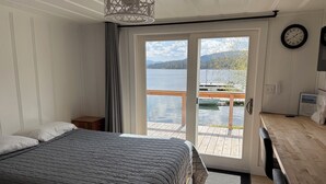 Een slaapkamer met een bed, een nachtkastje, een houten bureau, een stoel, een raam met uitzicht op een meer en een aanlegsteiger, en een klok aan de muur.