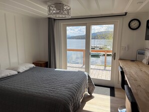 Um quarto com uma cama, uma mesa de cabeceira, uma escrivaninha de madeira, uma cadeira, uma janela com vista para um lago e um cais, e um relógio na parede.