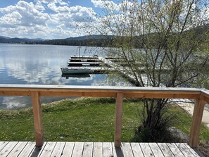 En træterrasse med udsigt over en rolig sø med fortøjede både, omgivet af grønt og fjerne bjerge.