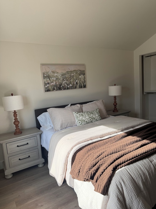 A bedroom with a bed, two bedside tables, a nightstand, a lamp, a painting, and a window.