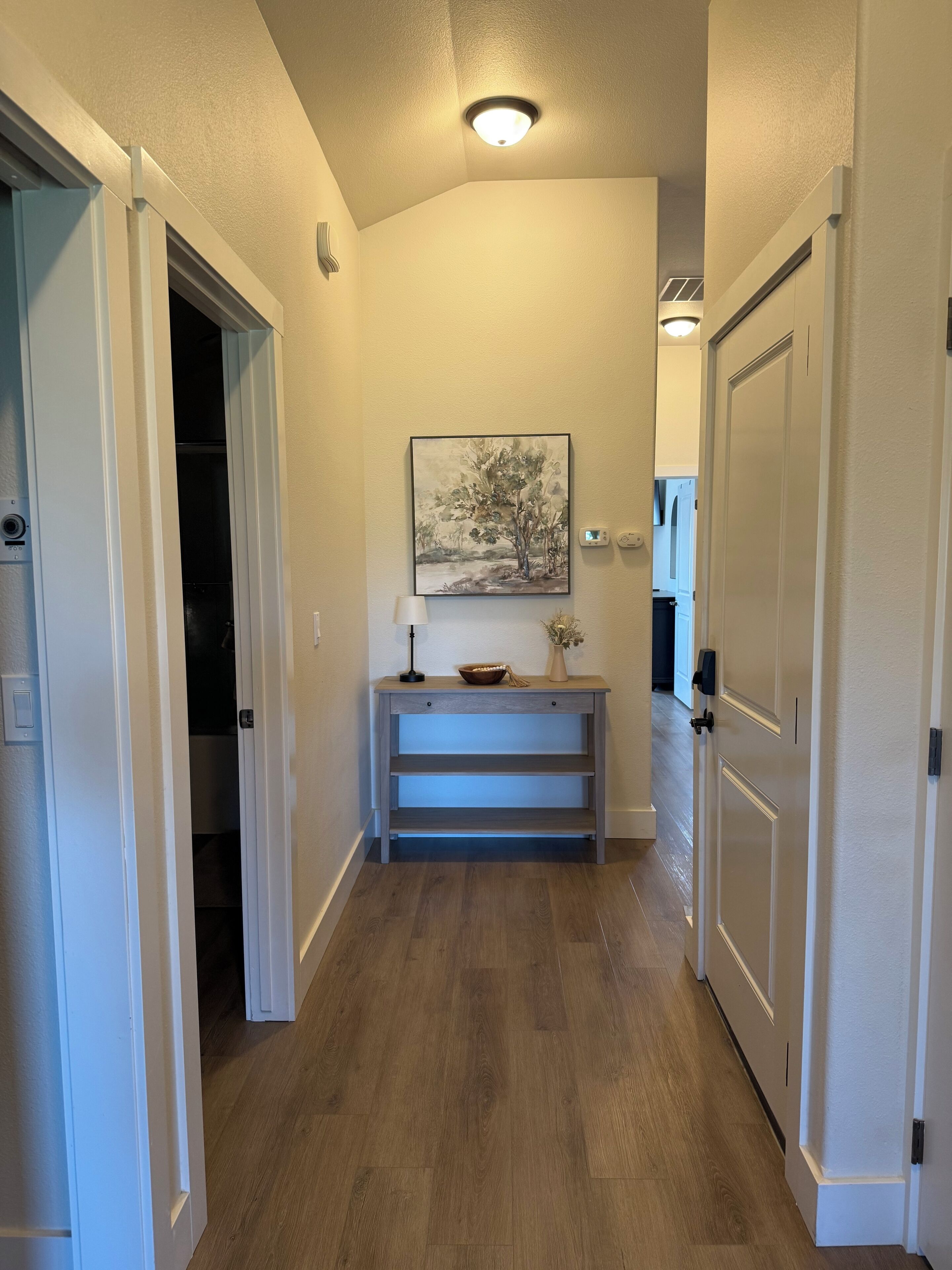 Un couloir avec un plancher en bois, un tableau accroché au mur, un bureau avec une lampe et une porte sur la droite.