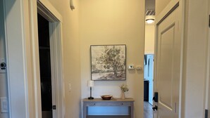 A hallway with a wooden floor, a painting on the wall, a desk with a lamp, and a door on the right.