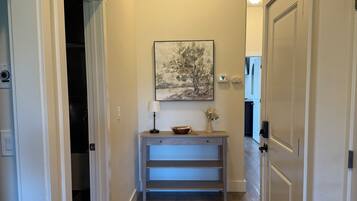 Un couloir avec un plancher en bois, un tableau accroché au mur, un bureau avec une lampe et une porte sur la droite.