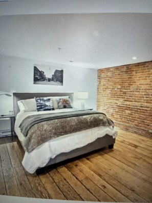 A modern bedroom with a wooden floor, a bed with a white and brown blanket, a nightstand, a lamp, and a brick wall. - Gorgeous 2-bedroom condo charming Stillwater overlooking St. Croix River (Stillwater)