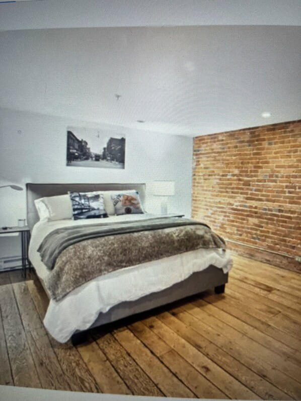 Ein modernes Schlafzimmer mit Holzboden, einem Bett mit weißer und brauner Decke, einem Nachttisch, einer Lampe und einer Ziegelwand.