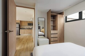 Ein Schlafzimmer mit einem Bett, einem Holzschrank, einem Spiegel und Blick auf eine Küche.