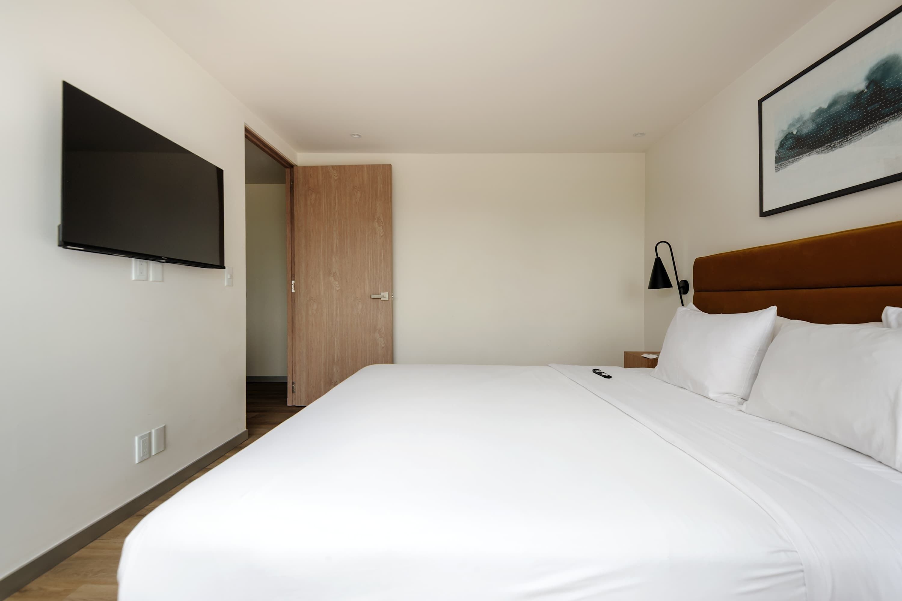 Habitació d'hotel amb un llit, una televisió penjada a la paret, una porta i una petita taula de nit amb una làmpada.