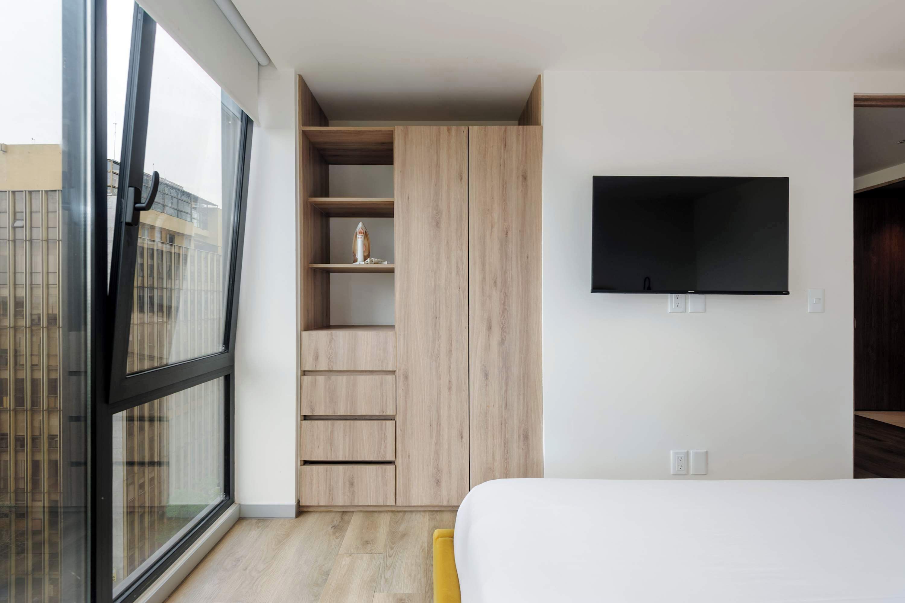 Une chambre avec une grande fenêtre, une armoire, un lit et un téléviseur fixé au mur.