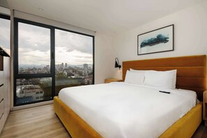 A hotel room with a large bed, a nightstand, a wall-mounted lamp, and a city view through floor-to-ceiling windows. - w* | Modern 2BR in Cuauhtemoc (Ciudad de México)