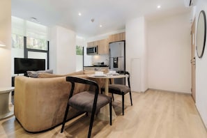 A modern living space with a sofa, dining table, chairs, a flat-screen TV, kitchen area with appliances, and large windows. - w* | Sophisticated 2BR in Cuauhtemoc w/ AC (Ciudad de México)